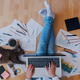 Top-down view of a person working on the floor, laptop on lap, surrounded by papers and pencils and a cat with a post-it note on her belly