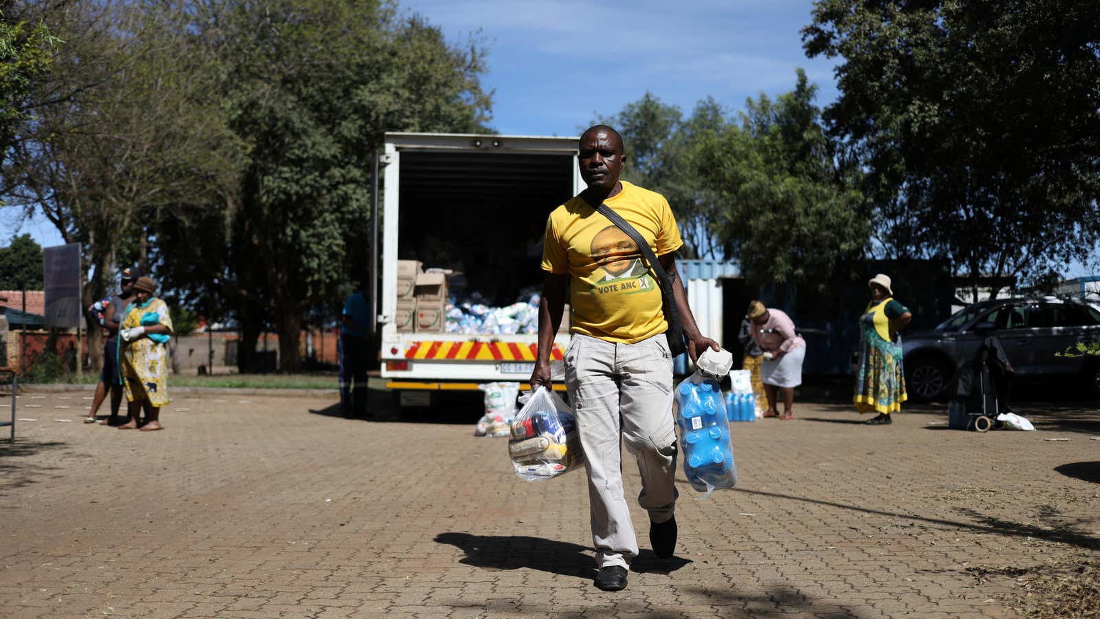 A man wearing a t-shirt with the face of South Africa’s president, Cyril Ramaphosa leaves as people distribute food parcels prepared by the local government…