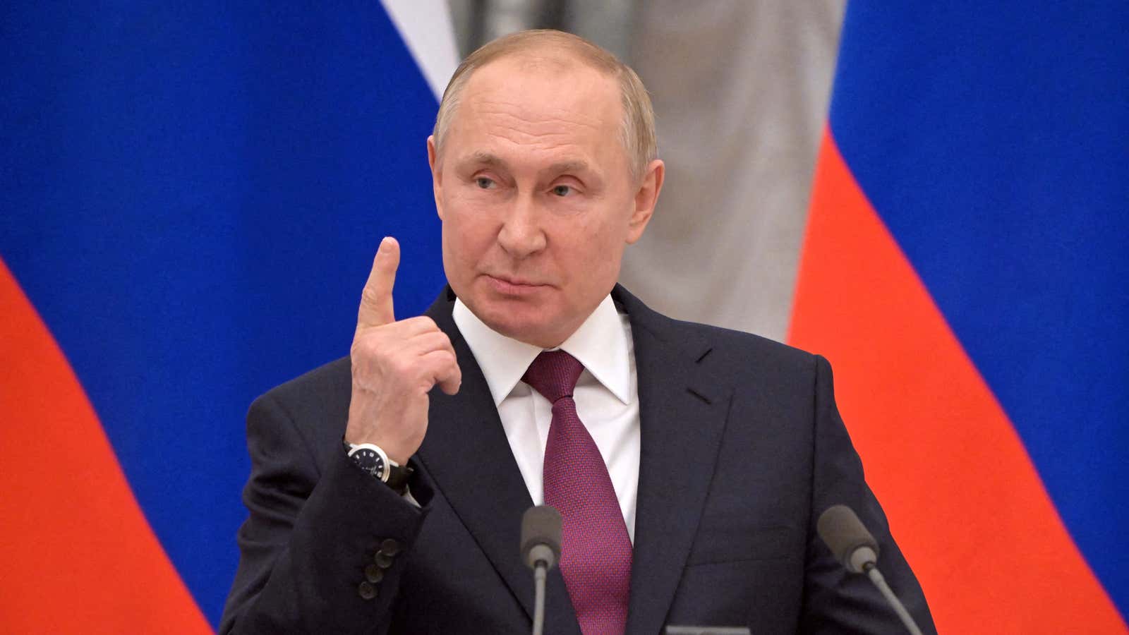 Russian president Vladimir Putin stands at a podium while raising the index finger on his right hand. Two Russian flags are displayed behind him.