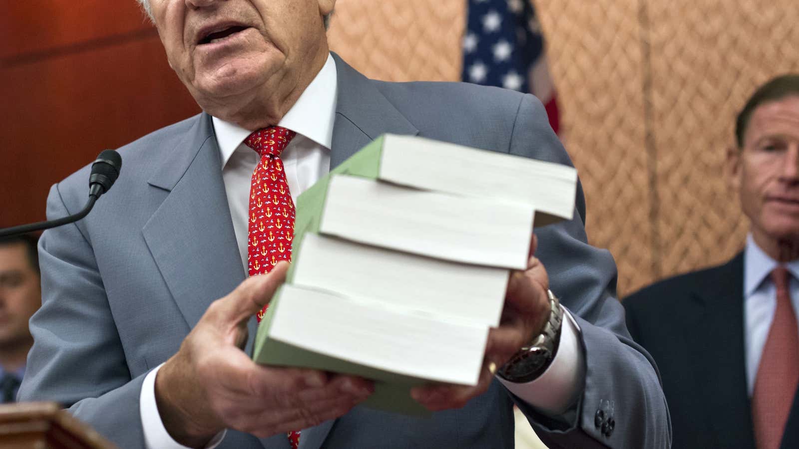 A senator holds up a four-volume report on malignant for-profit college practices.