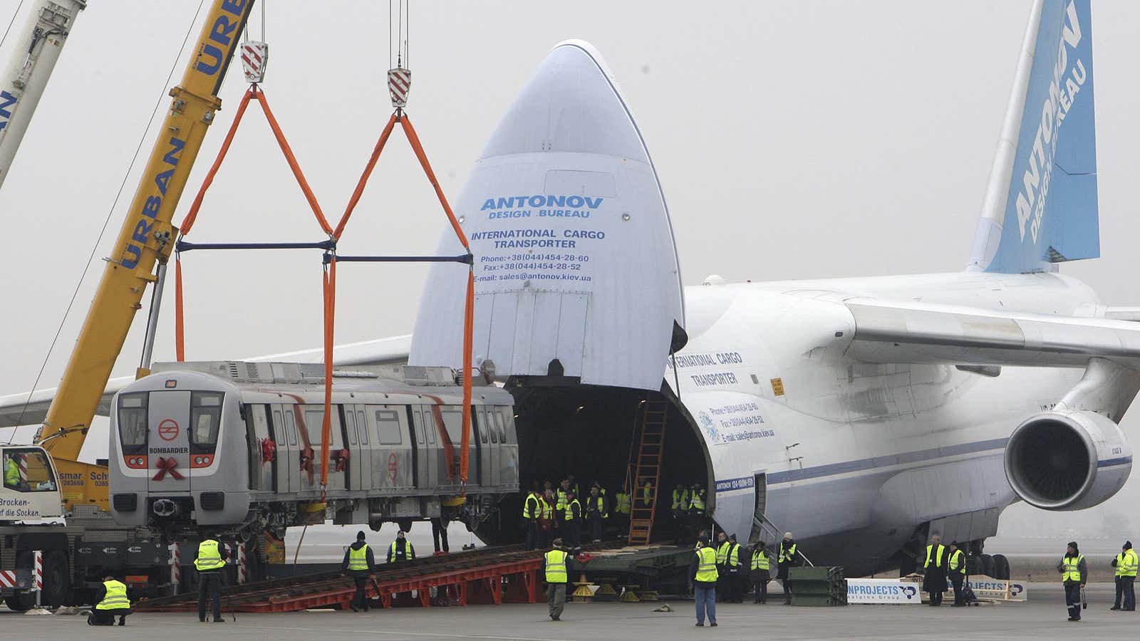 An An-124 sits on a runway. The plane’s entire nose lifts up on a hinge to reveal its cavernous cargo hold. A crane heaves a…