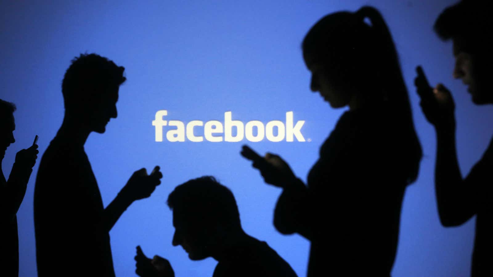 People are silhouetted as they pose with mobile devices in front of a screen projected with a Facebook logo, in this picture illustration taken in…