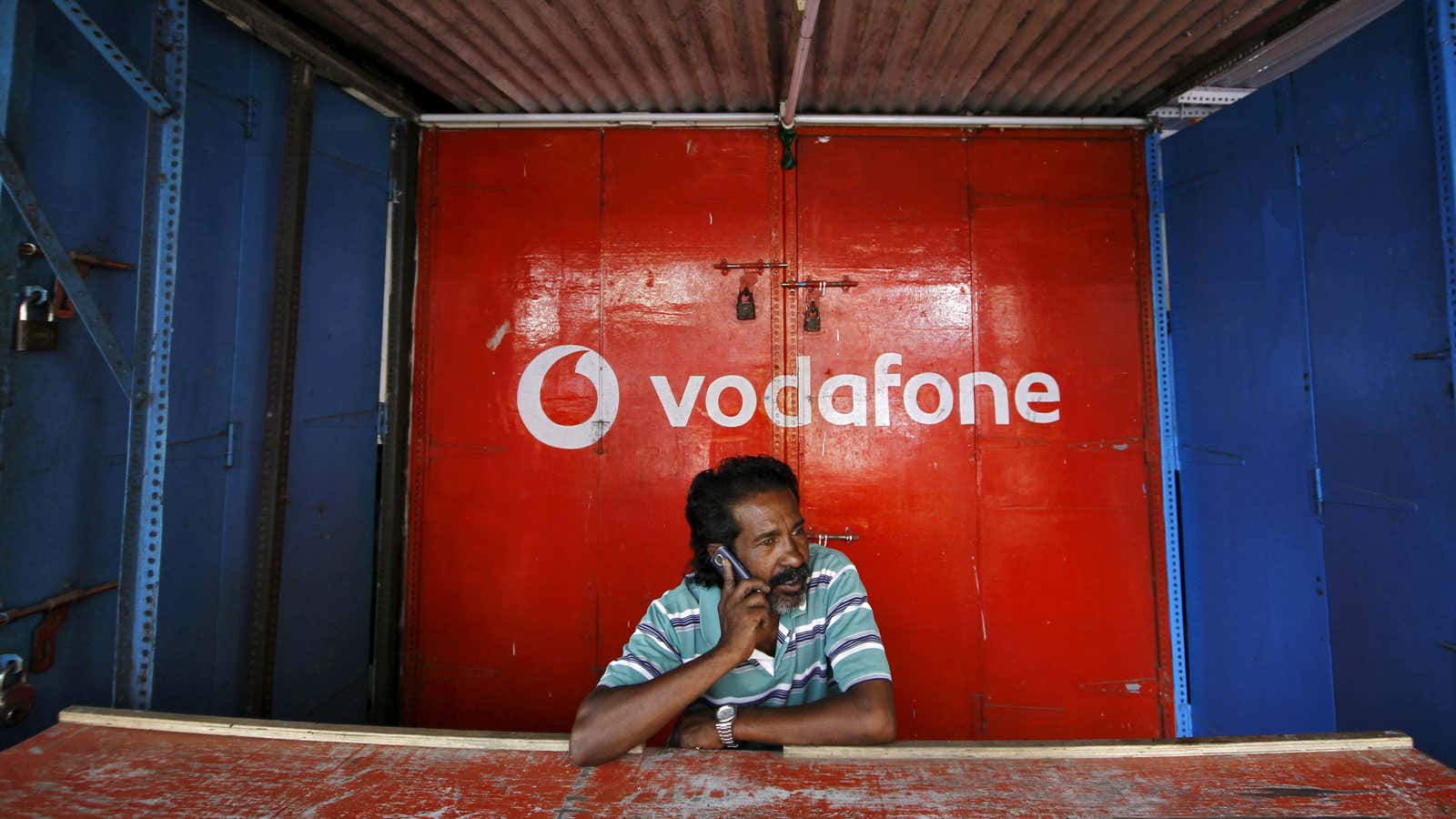 A retail shop owner speaks on his mobile phone outside his closed shop shutters painted with an advertisement for Vodafone at a market in the…
