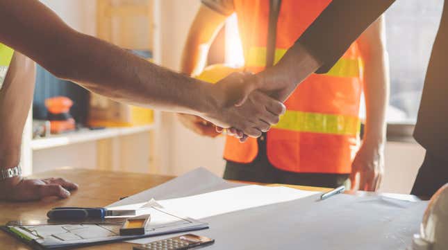 Photo of a contractor shaking hands with a client over a table spread with paper plans, tools, pens, and a calculator. Another contractor wearing a safety orange vest holds a yellow hardhat in the background.