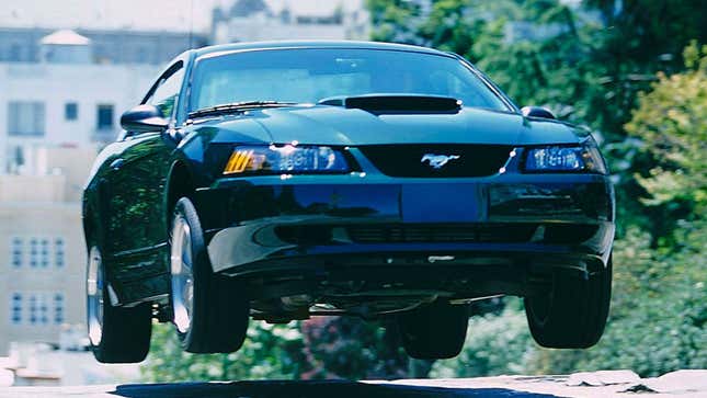 A photo of a dark green Ford Mustang coupe jumping over a bump in the road. 