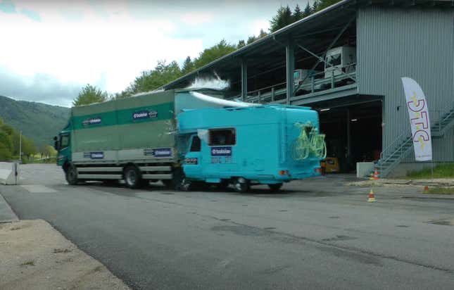Mini RV berwarna biru cerah menabrak bagian belakang sebuah truk komersial besar berwarna hijau.
