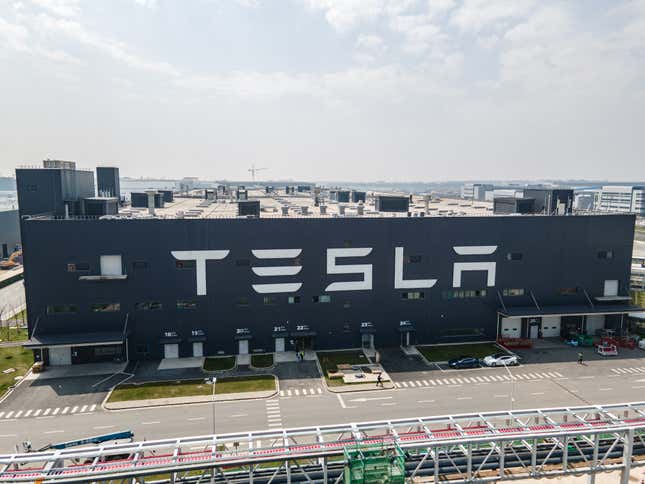 Tesla Shanghai Gigafactory is shown, a large, dark colored building with the word Tesla in white on one of its walls.