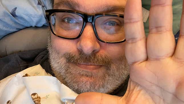 Mike Fahey of Kotaku says hi to the camera with his hand while lying in a bed.