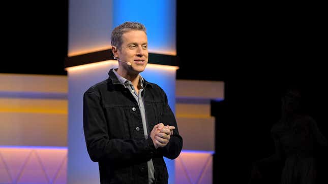 Geoff Keighley clasps his hands on stage as a zombie walks toward him in the background.
