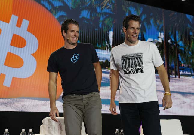 Tyler and Cameron Winklevoss stand on stage staring to the right with a giant bitcoin symbol blares on a screen behind them.