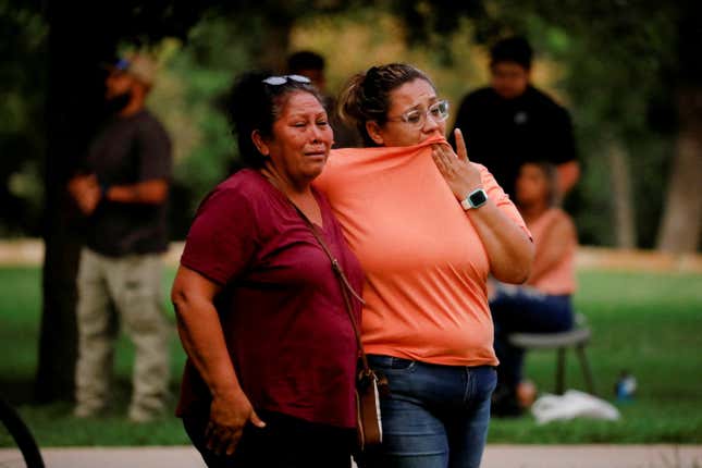 Two women, one in a dark red shirt, the other in orange, stand next to and console one another. They are visibly shaken and crying.