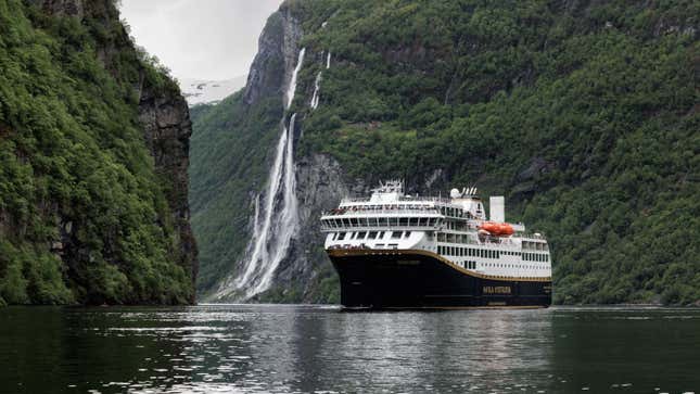 A small cruise ship floats peacefully in a fjord in front of a waterfall.