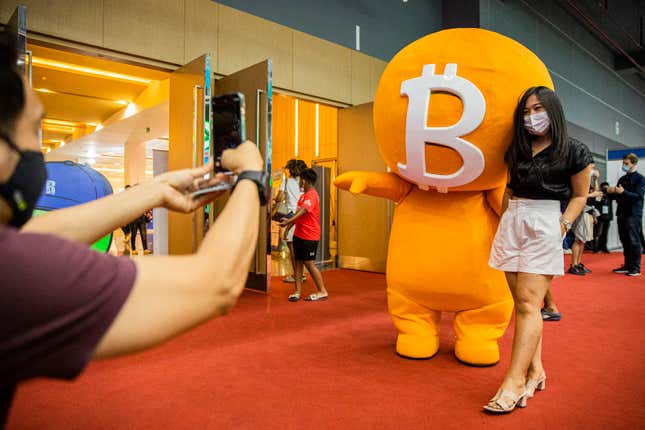 A man to the left takes a picture of a young woman standing alongside a large mascot bearing the bitcoin logo on its massive head.