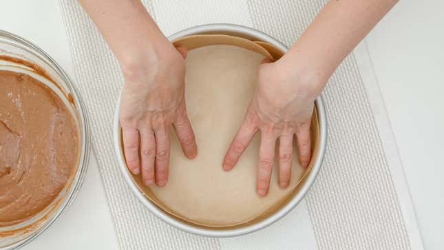 Hands lining round cake pan with parchment paper for baking