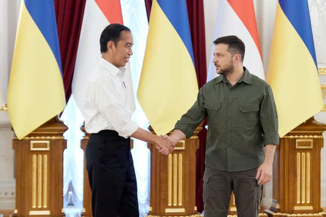 Indonesian leader Joko Widodo in a white dress shirt and black trousers shaking hands with Ukrainian president Volodymyr Zelenskyy in his trademark olive green, in front of some flags.