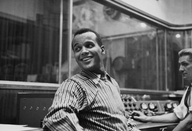 Harry Belafonte, wearing a striped shirt, in an unspecified recording studio, location unspecified, circa 1957. The sound engineer is visible working at the console to the right of the frame.