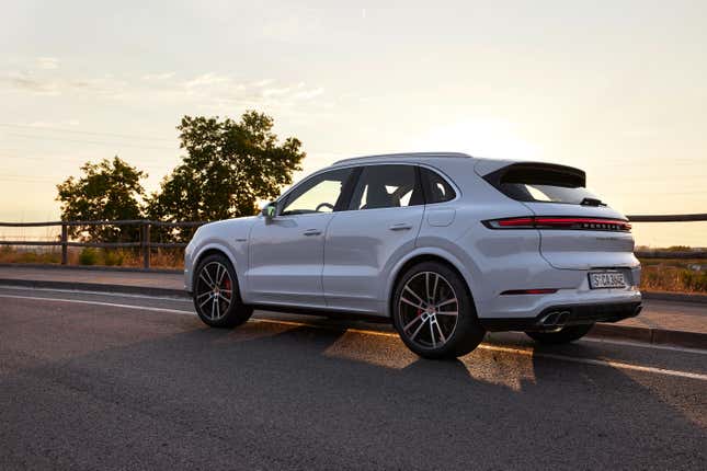 A white 2024 Porsche Cayenne Turbo E-Hybrid is parked on a road at sunset, rear three-quarter view