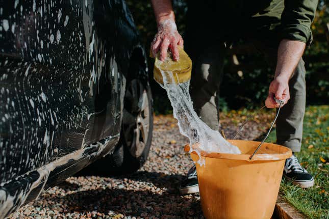 A man bends down to wash a car, using a sponge and bucket filled with soapy water. The sponge is saturated with water, creating a bubbly splash.