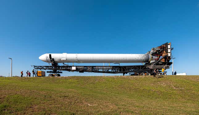 A view of the Terran 1 rocket on its side, being transported across Cape Canaveral. In the foreground is green grass, and the sky behind it is cloudless and blue.
