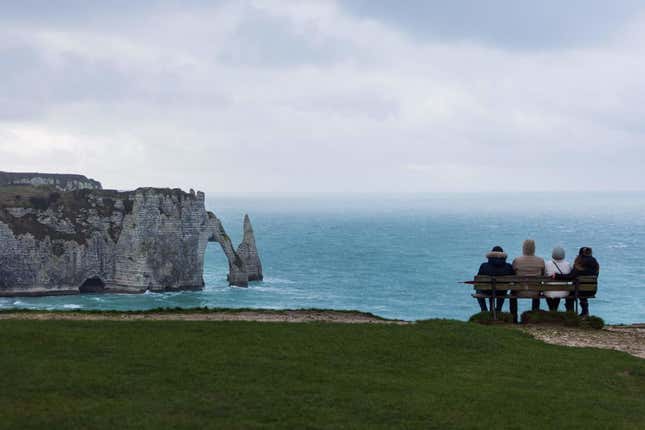 People sit on bench looking at a natural arch and the chalk cliffs in Etretat on January 4, 2022.
