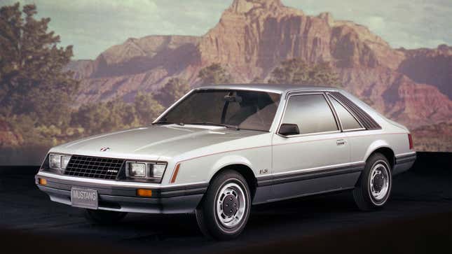 A photo of a silver Fox Body Ford Mustang with mountains in the background. 