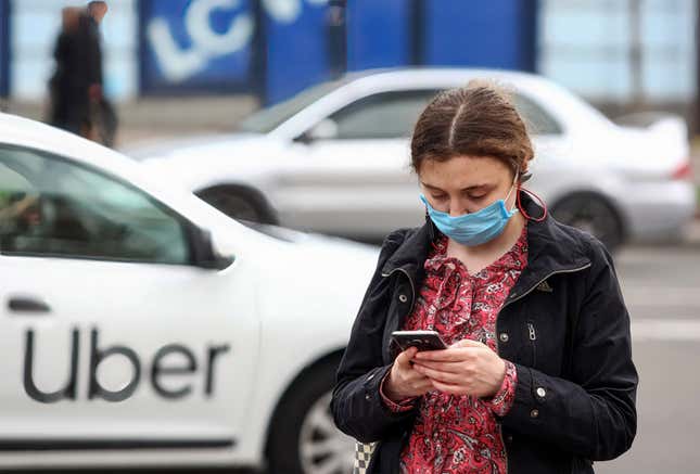 A woman wearing a facemask uses her phone as an Uber vehicle passes by in the background. 