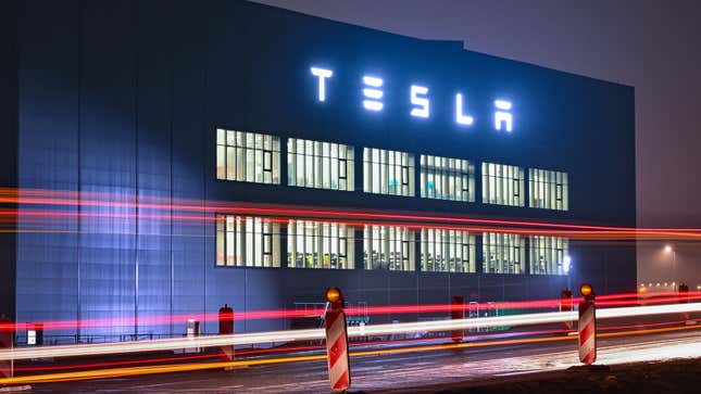 A photo of cars passing by a Tesla plant at night. 