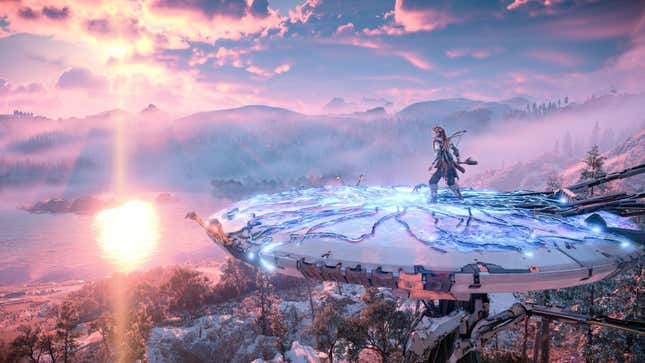 Aloy stands atop a Tallneck, in front of snowy hills, as the sun sets.