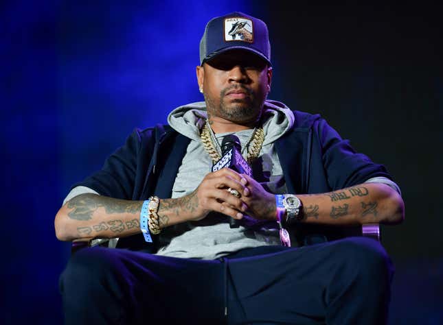 A Black man in a black baseball cap, gray hoodie, black jacket, and large gold chains around his neck and wrist holds a microphone while seated.