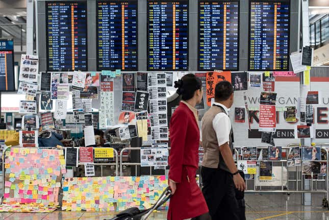 Flight attendants walk past an electric display board covered with notes and posters at Hong Kong’s airport on Aug. 13, 2019, the day after the airport closed due to protests.