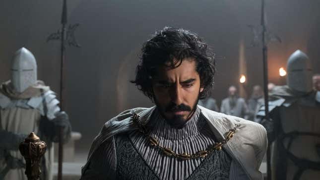 Dev Patel's Gawain in The Green Knight has his head bowed and is wearing several layers of grey with a gold chain holding his cloak. Two armor clad knights stand behind him on either side.