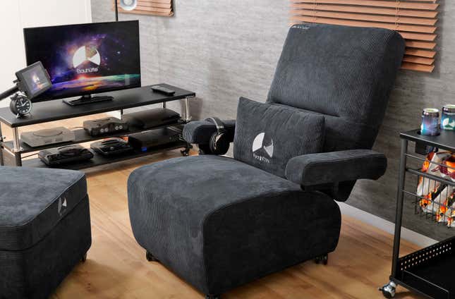The Bauhutte Gaming Sofa Deluxe in black parked next to a home entertainment center featuring several consoles.