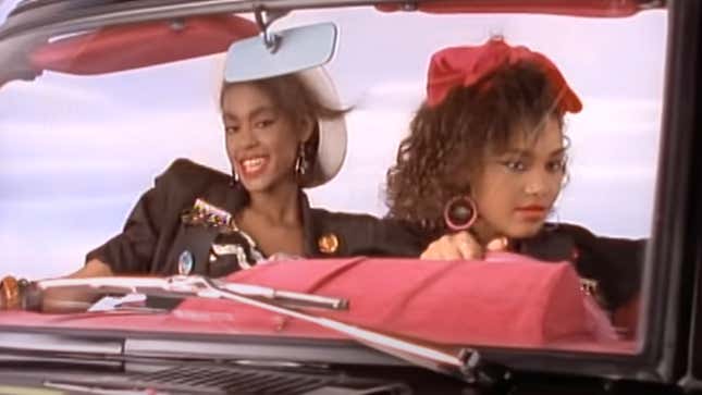 Two Black women in a car wearing 90s fashions and looking amazing