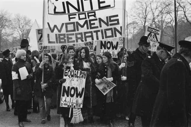 A black and white photograph of women protesting with signs. One reads "unite for women's liberation" and another reads "equal pay now." A few cops are visible next to the protestors.