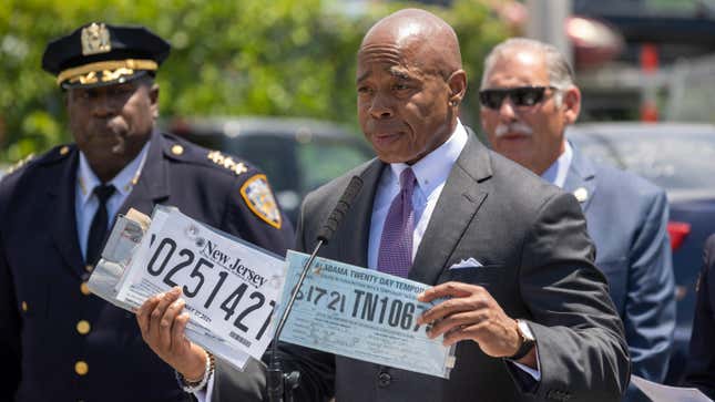 New York's Mayor holds up two examples of temporary paper plates used by drivers to dodge the law on a bright sunny July day. A high ranking police officer stands behind him and to the Mayor's right. On his left is a man in a suit wearing sunglasses.