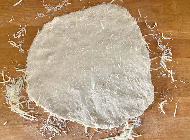 Shredded cheese on the counter with pizza dough on top. It's been stretched into a circle. Shredded cheese is pressed into the dough on both sides.