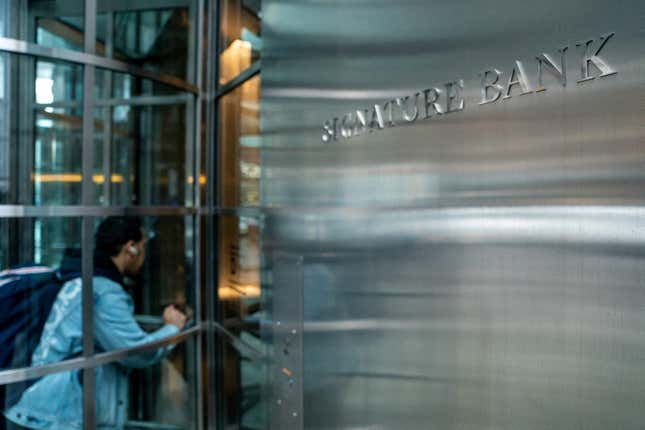 Signature Bank's metallic-facade headquarters. The name of the bank is mounted on the wall. A man in a light wash denim jacket enters the building through a clear glass revolving door.