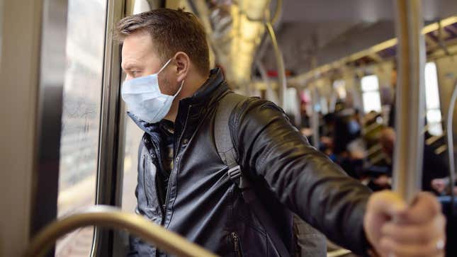 A man in a leather jacket and a mask stares pensively out the window while riding a train