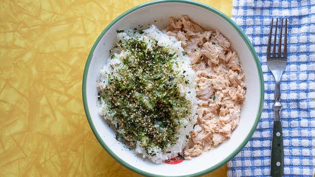 Top-down photo of a bowl of rice and tuna topped with furikake. The bowl sits on a yellow table with an abstract pattern and there's a blue-and-white checked napkin with a fork to its right.