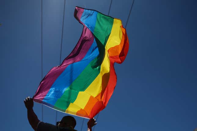 A person lifts a rainbow flag above their head, set against a blue sky.