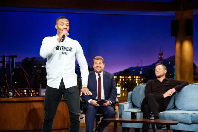 A Black man in black pants and a white Givenchy shirt raps into a microphone while two white men behind him smile and look on from a couch.