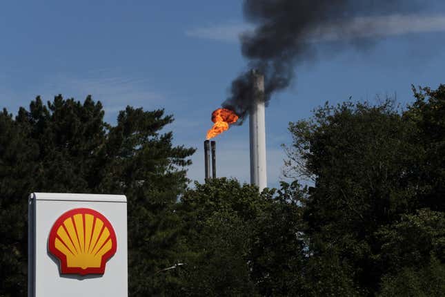 The sign of a Shell petrol station is seen in front of a burning pilot flame atop a flare stack
