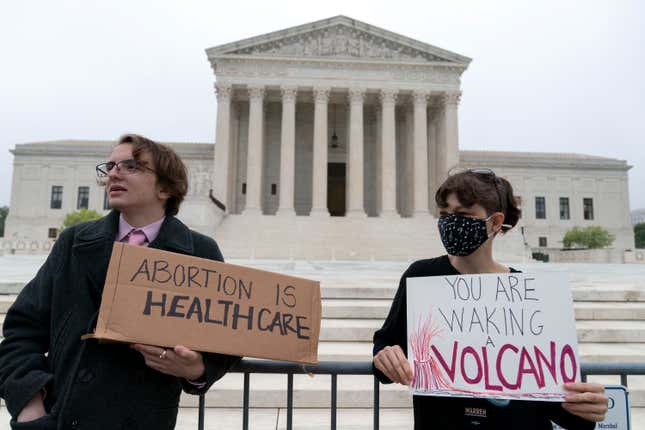 Two protestors stand outside the Supreme Court building holding signs. One says "abortion is healthcare," the other says "you are waking a volcano."