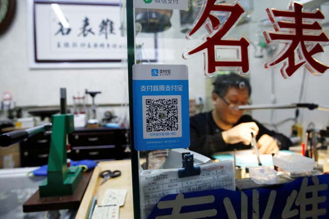 A blue-trimmed Alipay QR code is stuck to the glass pane outside of a watch repair shop. A man visible through the glass is working at a desk.