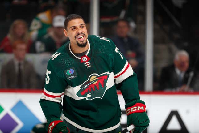 A Black man smiles in a green hockey uniform while holding his stick across his body on the ice.
