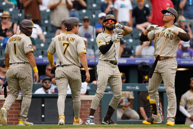 Two baseball players, in gray uniforms with brown stripes and helmets, walk back to the dugout while two of their teammates make celebratory gestures beside them.
