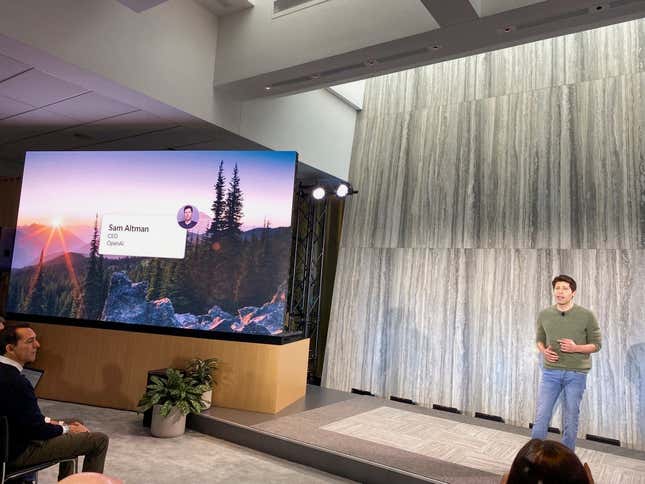 CEO of OpenAI Sam Altman speaks on a raised stage next to a presentation in a conference room.