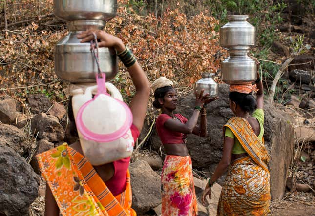 A woman helps another in carrying metal pitchers filled with water