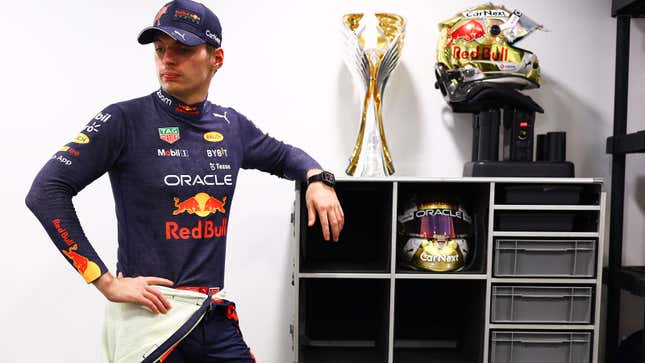 A photo of Max Verstappen in Red Bull gear leaning against a shelf with a trophy on top. 