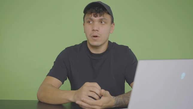 A young man sits in front of a green screen.
